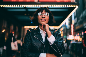 Millennial woman in trendy glasses with neon reflection looking up during evening sightseeing in...