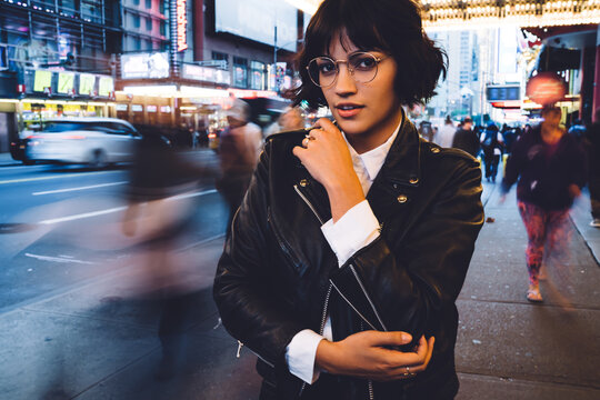 Half Length Portrait Of Stylish Hipster Girl In Cool Eyeglasses Standing Outdoors In Downtown With Time Lapse Of Crowd, Attractive Young Woman Dressed In Leather Jacket Looking At Camera On New York