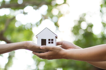 Hand giving home model to another person with green park background. metaphor house loan,  insurance and mortgage.