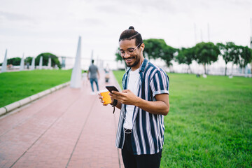 Cheerful dark skinned caucasian male meloman satisfied with music in earphones downloading songs to smartphone playlist via 4G connection, smiling african american man listening audio book on street