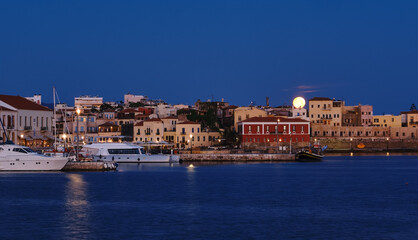 Fototapeta premium Full moon setting over Chania's Old Venetian Harbour. Night view of piers, boats and Maritime museum of Crete and houses over marina.