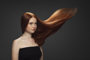 Fototapeta premium Youth. Beautiful model with long smooth, flying red hair isolated on dark grey studio background. Young caucasian girl with well-kept skin and hair blowing on air. Concept of salon care, beauty