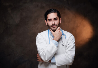 Adult and confident doctor with huge experience and stethoscope poses with hand under his chin in dark background.