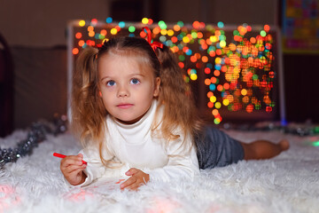 a little girl with two ponytails in a white sweater lies on a white rug and draws against the background of Christmas lights