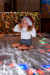 a little girl in a Santa hat sits near the Christmas lights