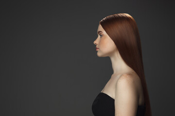 Glowing. Beautiful model with long smooth, flying red hair isolated on dark grey studio background. Young girl with well-kept skin and hair blowing on air. Concept of salon care, beauty, fashion.