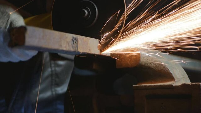 A locksmith with an angle grinder cuts off a piece of metal pipe. Sparks from cutting metal scatter like fireworks. The room is dark, only the lamp shines on the machine. 4k