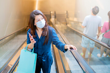 Fototapeta na wymiar Social distancing. Asian woman wearing a hygiene protective mask over her face, holding shopping bags and using mobile phone while standing on an escalator in a shopping center.