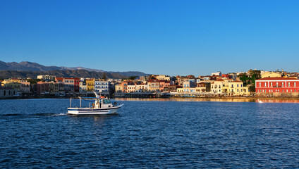 Fototapeta na wymiar White fishing boat passing by Old Venetian harbour quay and Maritime museum in Chania, Crete, Greece. Cretan hills and mountains. Early morning.