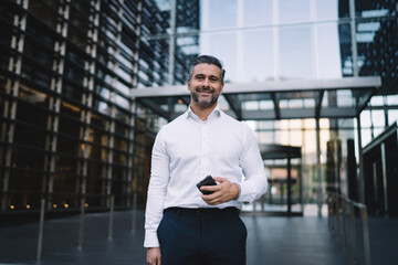 Half length portrait of happy male entrepreneur with modern cellphone technology in hand smiling at camera, successful mature businessman dressed in white shirt holding smartphone posing near office