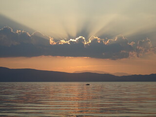 promienie zachodzącego słońca oświetlające chmury nad jeziorem, po którym płynie łódź © Agnieszka