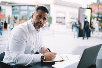 Obraz premium Portrait of mature entrepreneur in formal white shirt looking at camera during time for business planning, middle aged male employer working remotely in sidewalk cafe posing at table with netbook