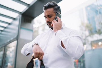 Middle aged male employee checking wrist watch time while waiting bus on stop calling to colleague for to warn about delay to business meeting,mature boss communicating via mobile during daily routine