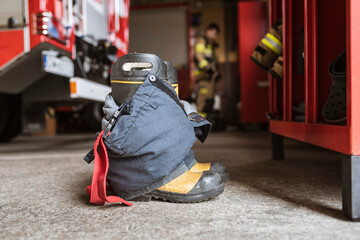 Firefighter's shoes and pants and a wardrobe cabinet, fire truck and fireman in the background.