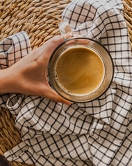 Mano sujetando un delicioso y cremoso café caliente servido en taza de cristal sobre una mantel de esparto y una servilleta estilo rústico