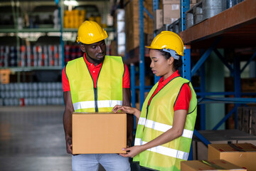 A Man worker holding a box or parcel and listen to a woman in the automotive parts warehouse distribution center. Both engineers people wear a safety helmet and vest. In background shelves with goods