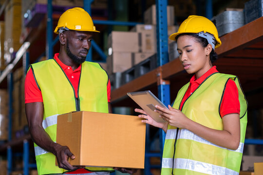 Woman worker using a tablet to check stock online and a man holding a parcel in the automotive parts warehouse distribution. Both engineers people wear safety helmet. In background shelves with goods