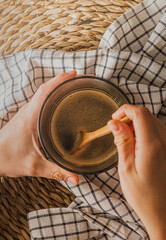 Manos sujetando y removiendo un delicioso y cremoso café caliente servido en taza de cristal sobre una mantel de esparto y una servilleta estilo rústico