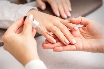 Tuinposter Hand of manicurist pours oil by pipette to cuticle of nails of young woman in beauty salon. French manicure © okskukuruza