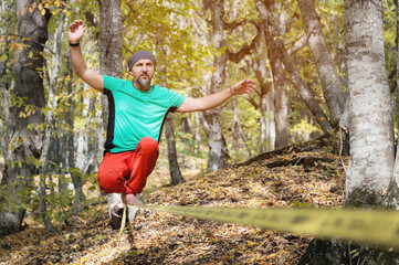Naklejka na ściany i meble A bearded man in age balances while sitting on a taut slackline in the autumn forest. Outdoor Leisure