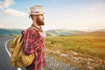 Young male tourist hitchhiker in a hat shorts with a backpack walks along a country asphalt road against the backdrop of mountains at sunset