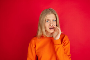 Shocked whisering secret. Portrait of young caucasian woman isolated on red studio background with copyspace. Beautiful female model. Concept of human emotions, facial expression, sales, ad, youth.