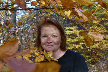 Rothaarige Frau Anfang 50 im gelben Herbstlaub hält ihr Gesicht in die Sonne, lächelt, gelbe und...