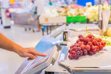The seller is weighing grapes and points the fingers on the screen electronic scales in supermarket.