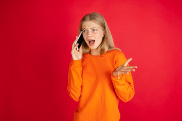 Shocked talking phone. Portrait of young caucasian woman isolated on red studio background with copyspace. Beautiful female model. Concept of human emotions, facial expression, sales, ad, youth.