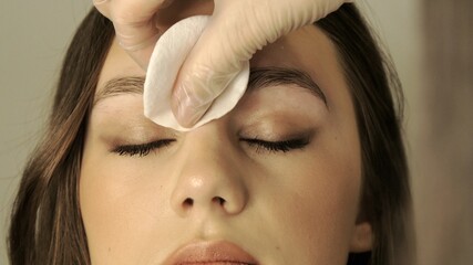 Cosmetologist's hands wiping eyebrows using cotton pads. Beautician rubs client's eyebrows with a cotton pad. Beautician in gloves is doing permanent makeup to girl. Eyebrow correction. 