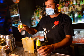 young bartender with face mask pouring cocktail inside bar. hospitality sector has been badly affected by the corona virus crisis