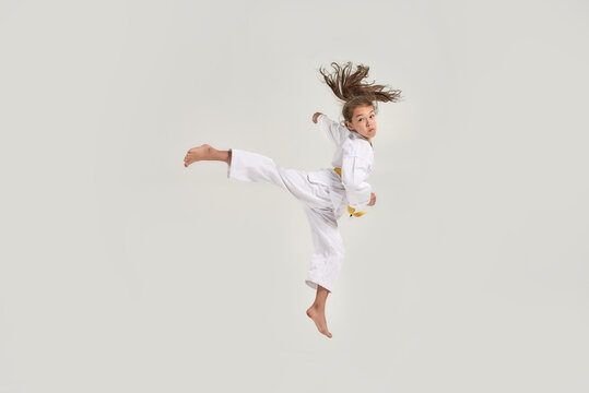 Full length shot of little karate girl in white kimono with a yellow sash exercising and fighting, doing martial arts, jumping isolated over white background