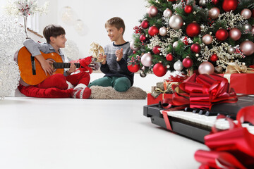 merry christmas and happy holidays, children play guitar and tambourine near the christmas tree with wrapped gift packages and musical instruments, at home in the living 
room sitting on the floor