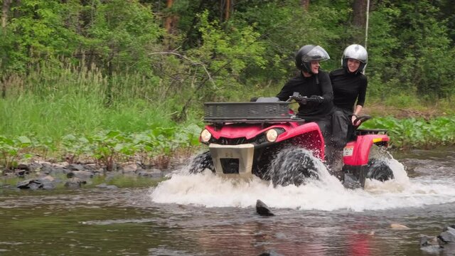 Slow motion tracking of cheerful man and woman in helmets riding red quad bike through river in forest