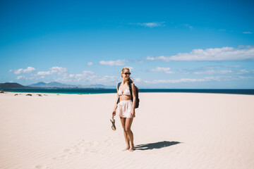 Happy carefree female tourist with backpack standing at nature horizon landscape and smiling outdoors, young cheerful woman solo traveller enjoying getaway on picturesque beach with white sands