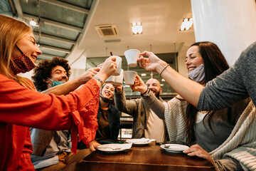 People with face mask drinking at coffee house - Young friends celebrating toasting with cappuccino cup at restaurant - New normal lifestyle concept with happy guys and girls at cafe bar
