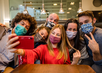 Group of friends covered by facemask at the restaurant - Young happy people celebrating taking a selfie with smartphone - People, technology and new normal lifestyle concept.