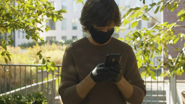 Portrait of hipster in black protective mask and gloves taking photos by smartphone in the park, at autumn sunny day. Steadicam shot