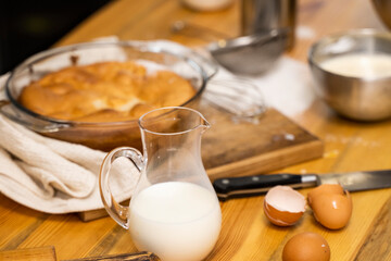 Tasty apple pie on a oaken wood table with a ingredients. Cooking a delicious apple pie on a kitchen in a loft style. Milk, eggs, sugar and a baked pie on a table. Home kitchen.