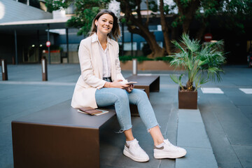 Portrait of carefree female student dressed in casual clothing sitting at urbanity and using mobile application for listening radio music, Caucasian hipster girl smiling at camera enjoying podcast