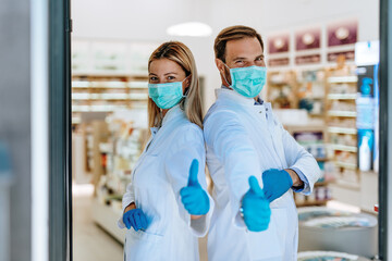 Male and female pharmacists with protective mask on their faces working at pharmacy. They are showing thumbs up. Medical healthcare concept.