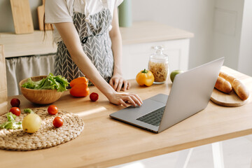 Fototapeta na wymiar Young caucasian woman in apron use laptop computer in the modern kitchen, preparing salad, read recipe.