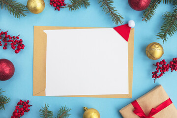 Christmas greeting card or New year Goals concept. Empty paper blank with envelope, spruce tree branches, gift box and festive decor on light blue background. Top view, flat lay, copy space, mock up