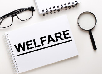 The word WELFARE is written in a white notebook on a light background near the notebook, black-framed glasses and a magnifying glass.