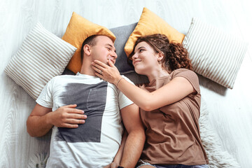 Obraz na płótnie Canvas Young lovely couple newlyweds lying on the floor with pillows and look at each other, romantic atmosphere. Calm after child go sleep, husband and wife relaxing, chilling indoor. 