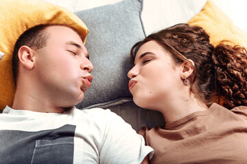 Young lovely couple newlyweds lying on the floor with pillows and look at each other, making faces grimaces, romantic atmosphere. Husband and wife relaxing, chilling indoor. 