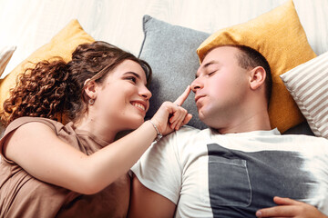 Young lovely couple newlyweds lying on the floor with pillows and look at each other, making faces grimaces, romantic atmosphere. Husband and wife relaxing, chilling indoor. 