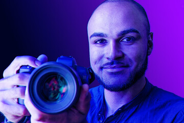Black man with camera standing against purple background in neon light
