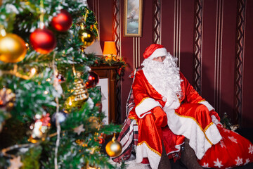 Portrait of Santa Claus - with a white beard, Santa's hat, with gifts near the Christmas tree and fireplace on an armchair on Christmas Eve and New Year.