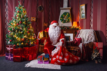 Portrait of Santa Claus - with a white beard, Santa's hat, with gifts near the Christmas tree and fireplace on an armchair on Christmas Eve and New Year.